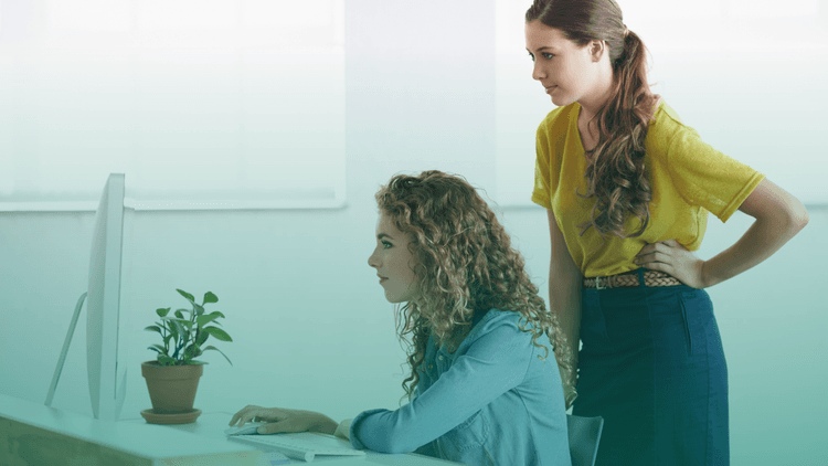 Duas mulheres analisam informações em um computador de mesa em ambiente corporativo, transmitindo foco e atenção ao realizar uma pesquisa ou verificação de dados.