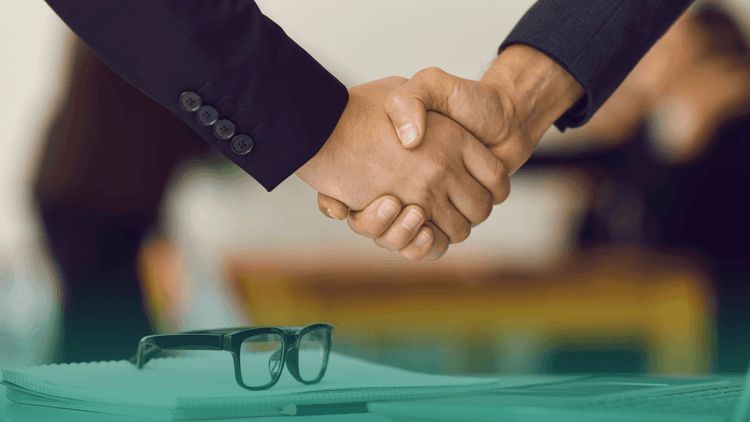 Duas pessoas apertando as mãos em um ambiente corporativo, simbolizando a formalização de uma contratação. Em primeiro plano, um caderno e um par de óculos sobre a mesa.